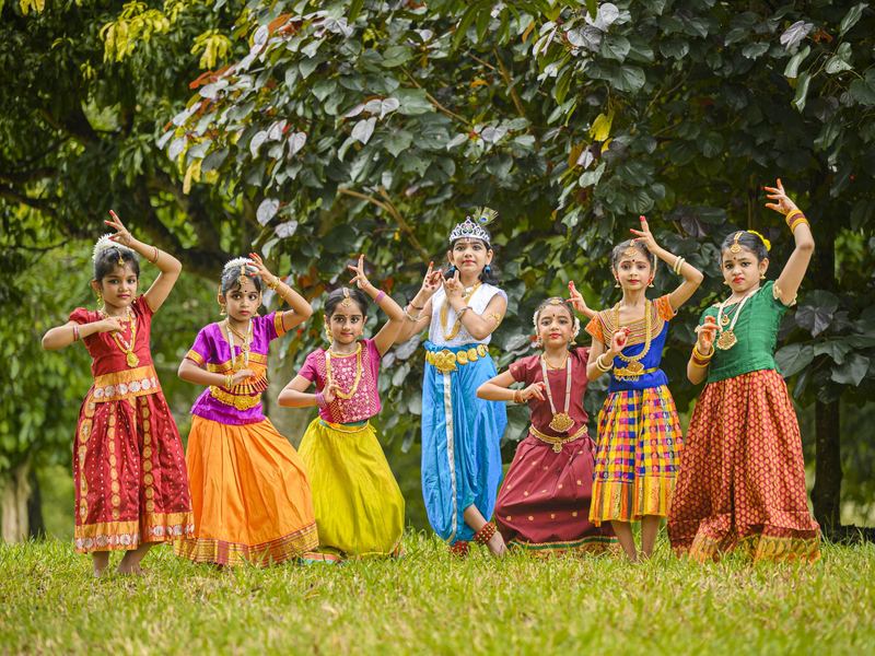 Seven young Bharatanatyam students in colourful costumes posing together on a green lawn at Konjum Salangai 2020